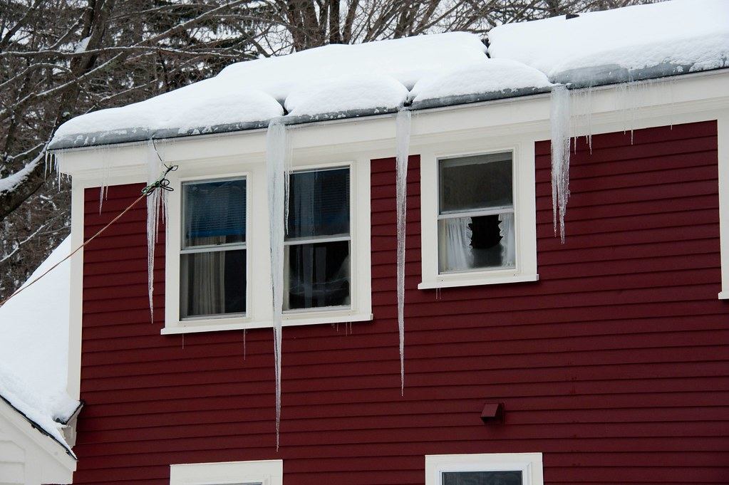Snow-covered roof edge with long icicles hanging over the windows of a red house